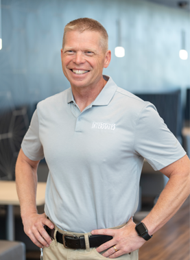 Scott Peterson, CEO of Interstates, smiling and standing with hands on hips in a modern office space. He is wearing a light gray Interstates-branded polo shirt and khaki pants, with a smartwatch on his wrist.